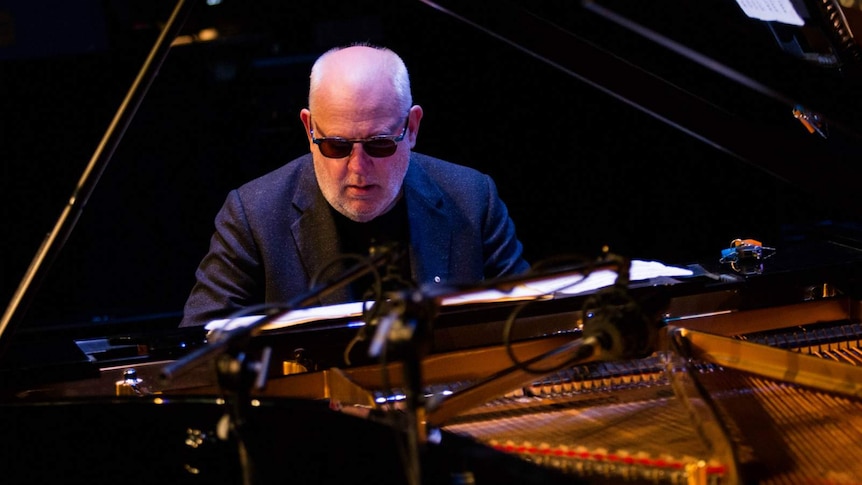 Paul Grabowsky sits at a piano, wearing glasses and performing
