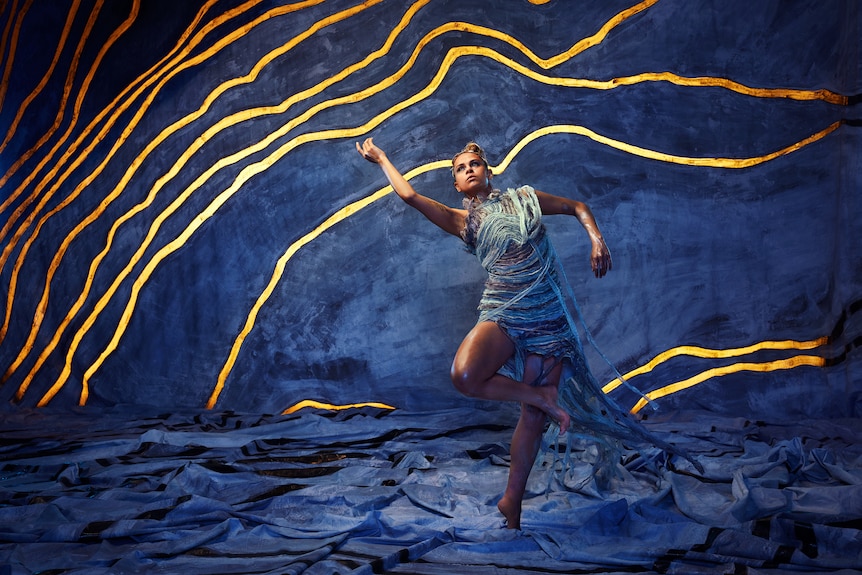 A First Nations female dancer wears blue and white embroidered costume mid-choreography on a vivid blue set cut with gold lines.
