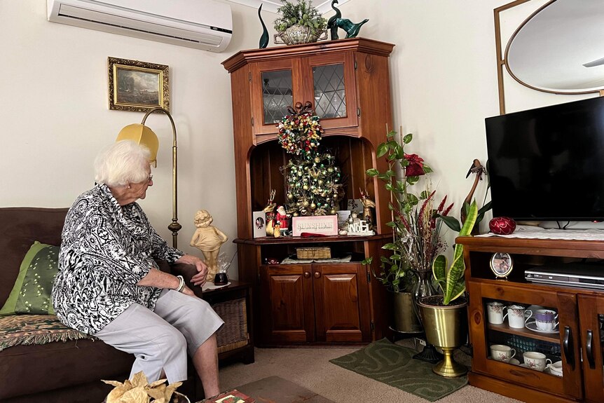An elderly woman in a black and white frock and grey pants looks at a small Christmas tree on a corner cabinet.