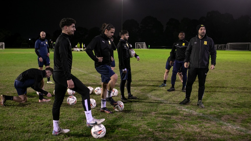 A number of football players, wearing black stand on a pitch at night. Some have soccer balls at their feet.