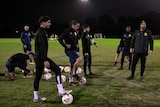 A number of football players, wearing black stand on a pitch at night. Some have soccer balls at their feet.