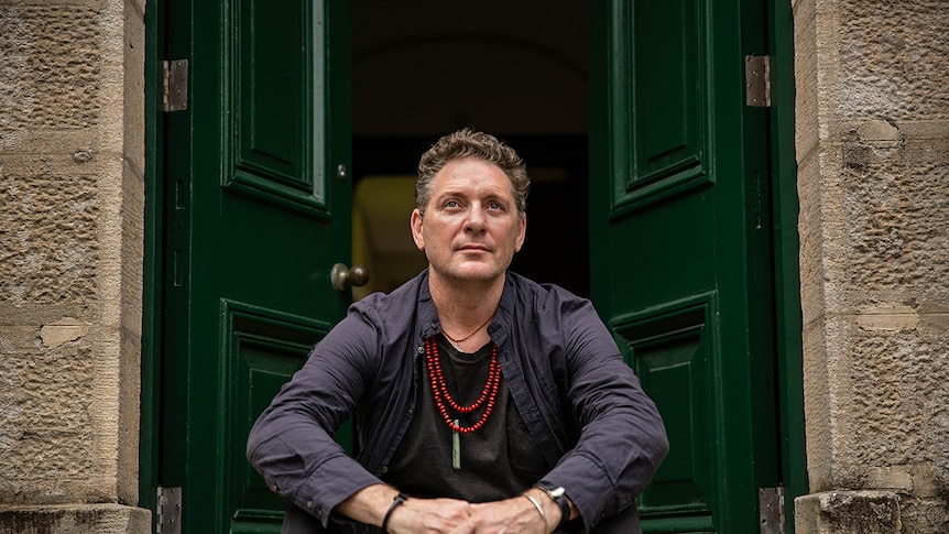 Colour photo of artist Brook Andrew sitting on the doorstep of a sandstone building at the National Art School in Sydney.