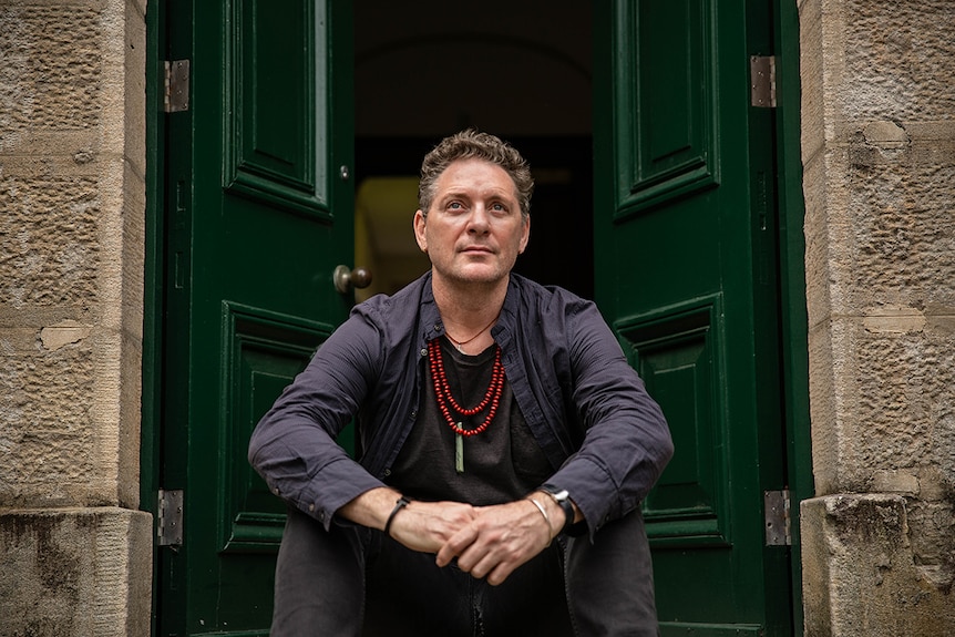 Colour photo of artist Andrew Brook sitting on the doorstep of a sandstone building at the National Art School in Sydney.