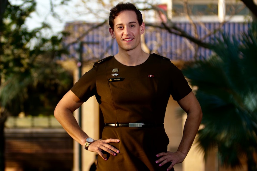 Smiling person, short brown hair, hands on hip, wears Australian Army uniform dress. Sunshine, blurred house, greenery behind.