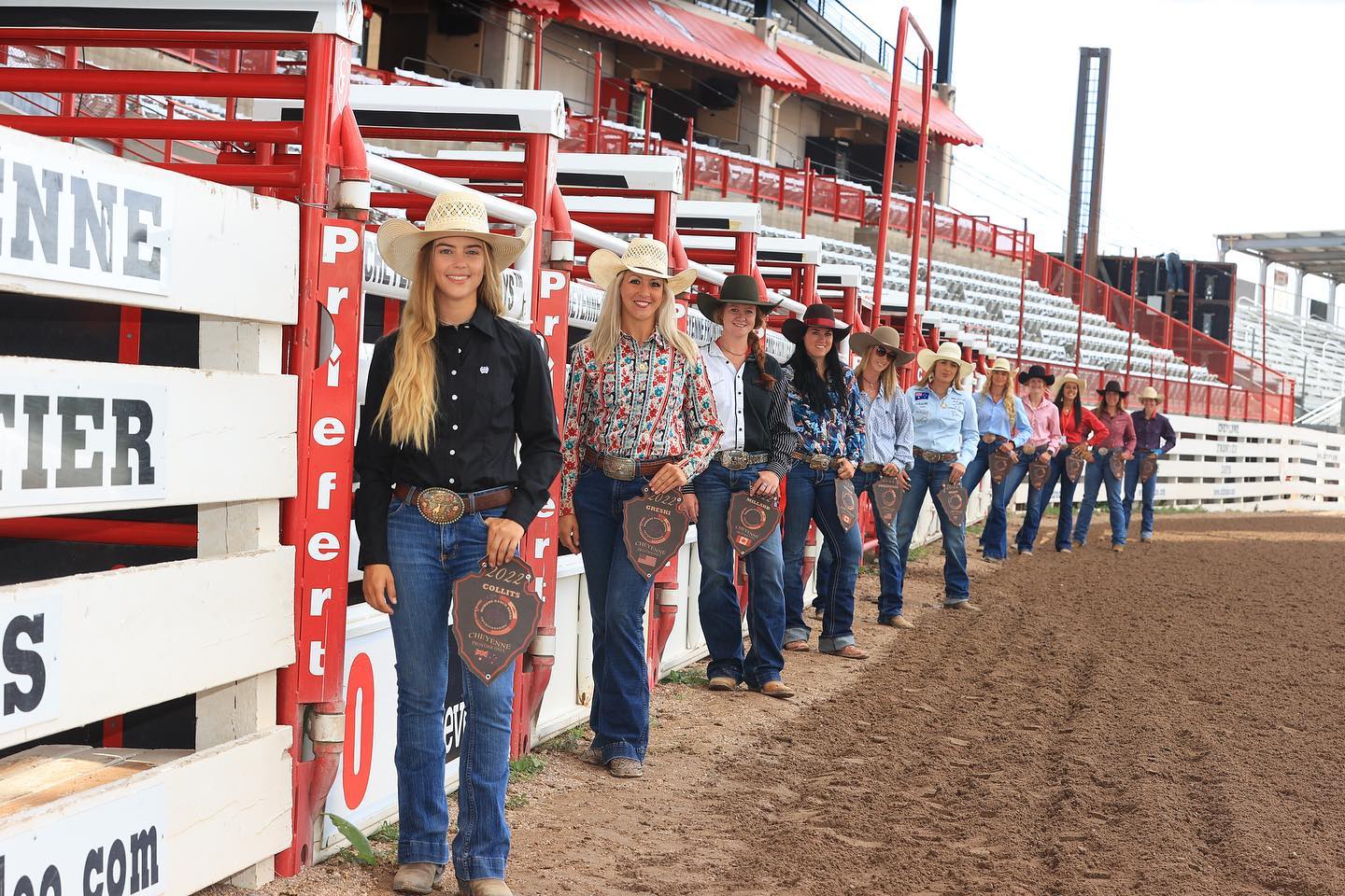 Eleven brightly dressed women in cowgirl hats stand along a row of horse chutes at a rodeo ground.