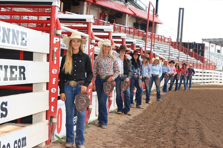 Eleven brightly dressed women in cowgirl hats stand along a row of horse chutes at a rodeo ground.