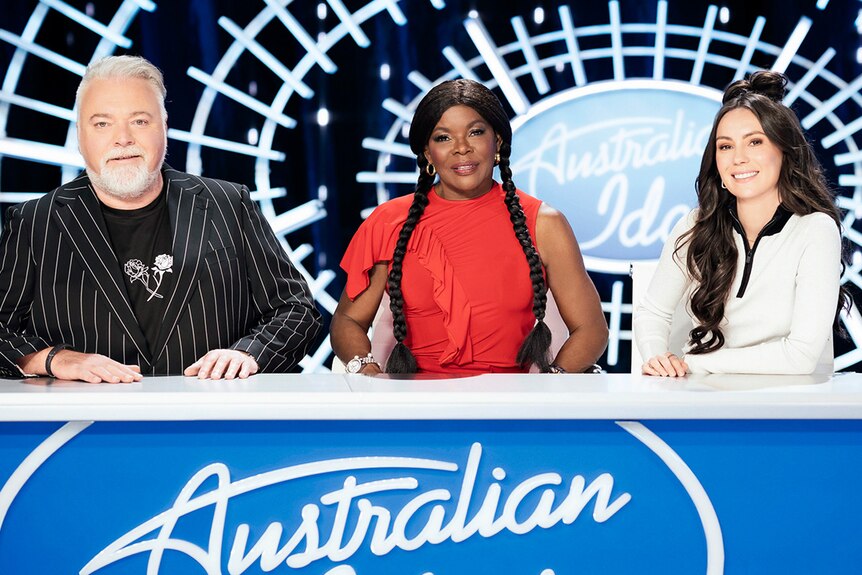 Kyle Sandilands, Marcia Hines and Amy Shark behind a desk with Australian Idol written on it. 