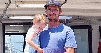 A photo of a man in a blue t-shirt and baseball cap holding a young boy on a boat.