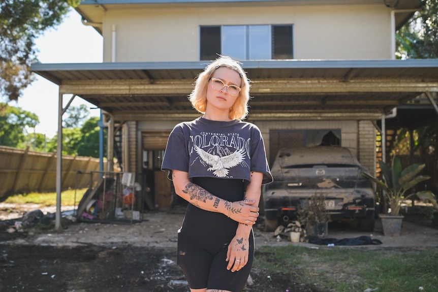 A woman with tattoos standing outside a wrecked house.