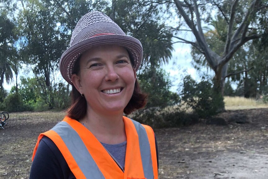 a woman in an orange vest smiles at the camera with bushland behind her