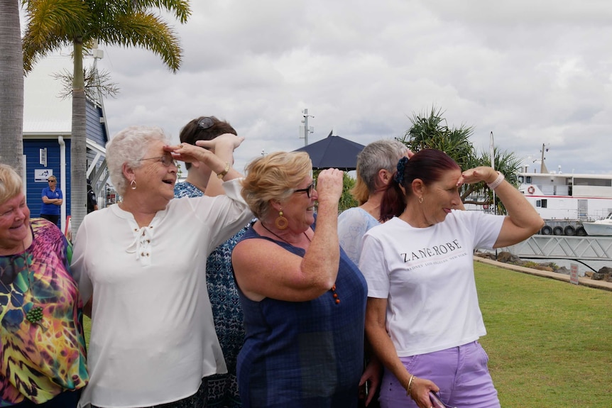 A group of six women looking out to sea, hands raised over their eyes, laughing.