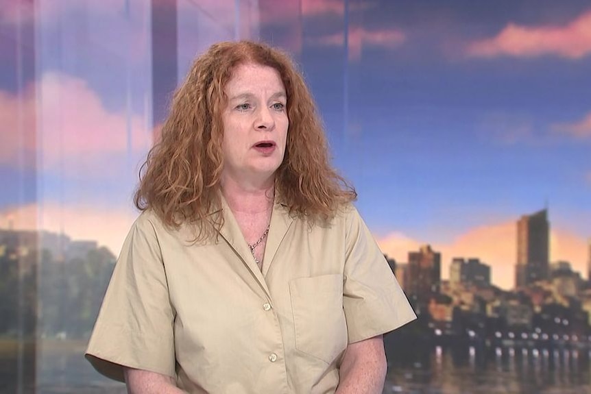 Correspondent sits in studio infront of a city skyline wearing a beige button up shirt and flowing auburn hair