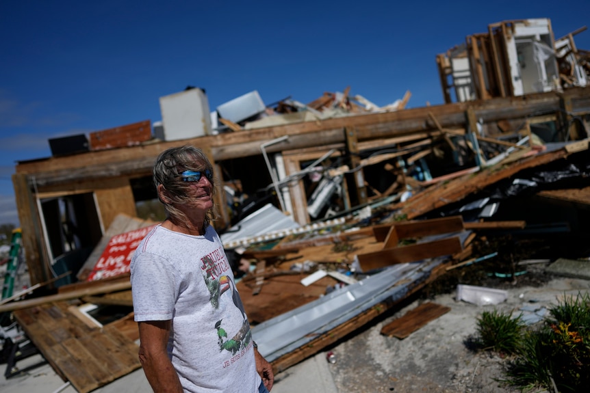 A man with windblown hair in a white T-shirt and sunglasses stands next to a destroyed building in bright sunlight.