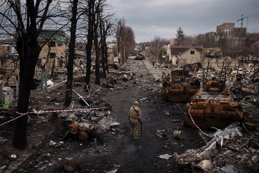 A soldier walks along a road littered with war wreckage, with bits of homes, vechiles and other structures strewn everywhere.