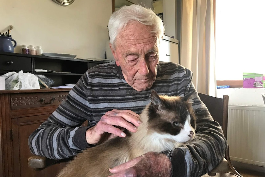 David Goodall sits inside cuddling a cat perched on his lap.