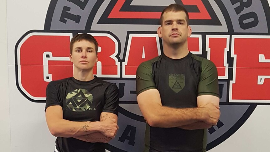 Two people stand in a Jiu Jitsu facility, facing the camera.