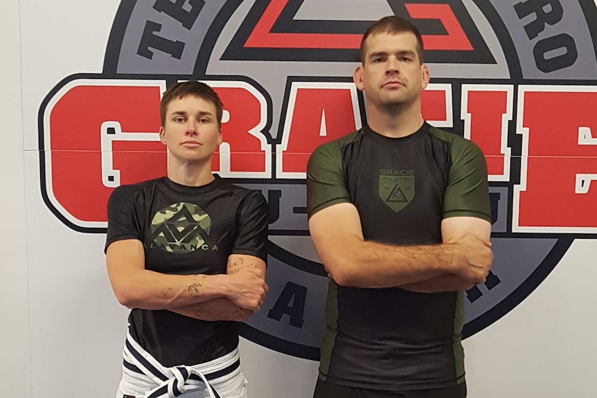 Two people stand in a Jiu Jitsu facility, facing the camera.