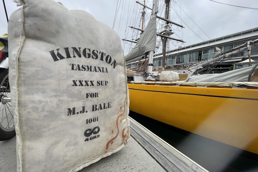 Bale of merino wool in a hessian bag in front of the yellow wooden boat it's about to be loaded onto at the Hobart dock.