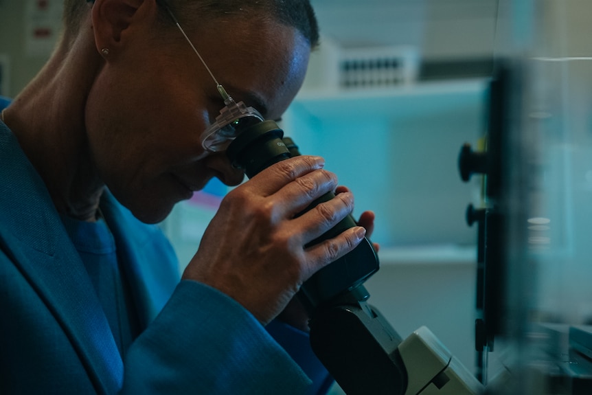 Female doctor wearing blue shirt and jacket looking through a microscope in a lab.