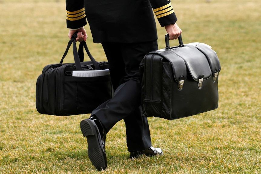 A military aide walks across a grassy area carrying two briefcases.