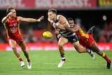 An AFLW player leans almost full-length as he wraps his arms around an opponent, who grimaces as the ball falls clear.