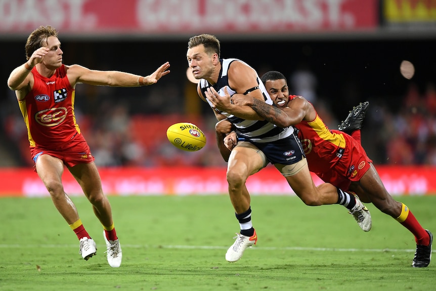 An AFLW player leans almost full-length as he wraps his arms around an opponent, who grimaces as the ball falls clear.