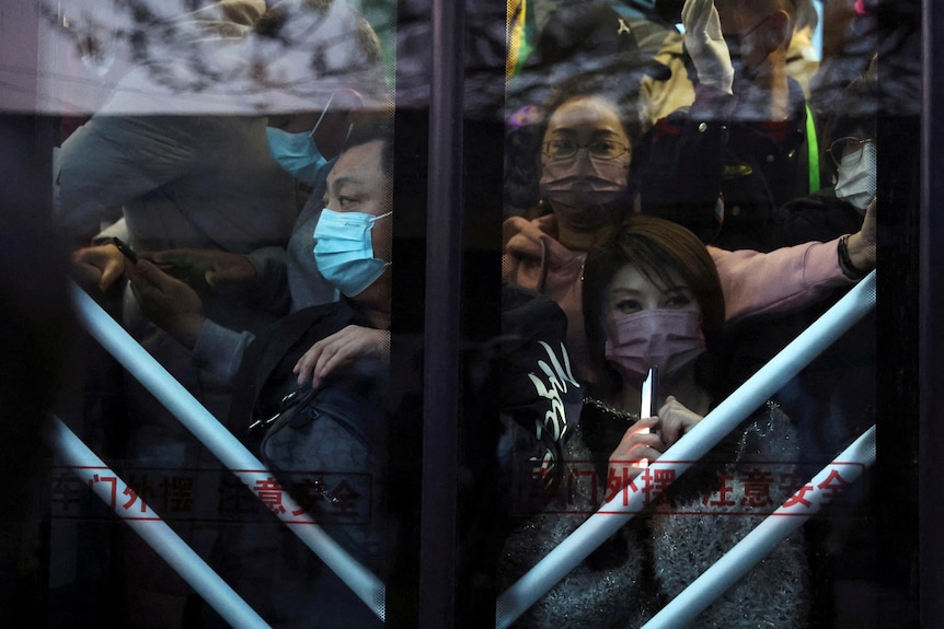 The glass doors of a train, through which you can see a packed crowd of commuters, all in masks 