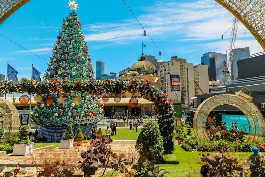 A Christmas tree, baubles and golden archways decorate Federation Square, under a blue sunny sky.