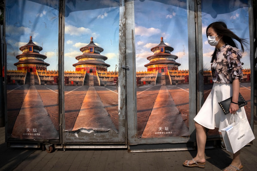 A woman with a shopping bag in a mask walking past a shop decorated with images of thr Temple of Heaven in Beijing.