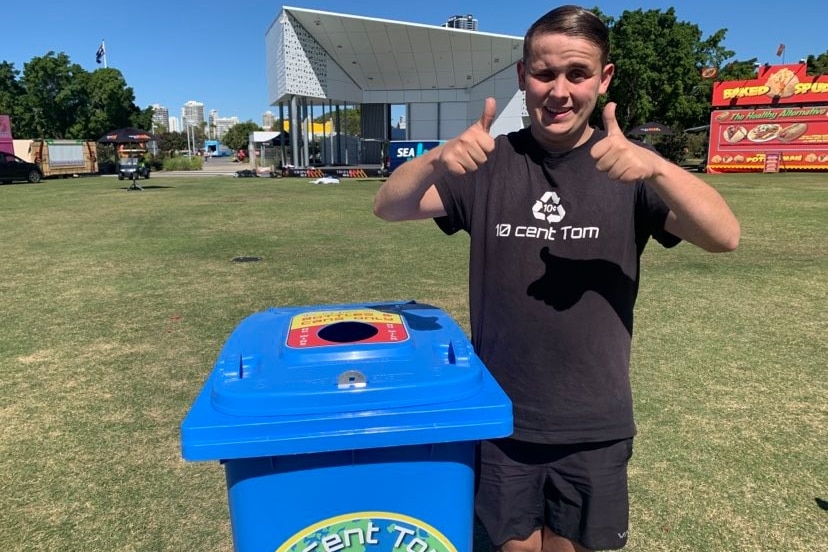 A teenager stands beside a blue recycling bin in the showground with his thumbs up. He's wearing a 10-Cent Tom T-shirt