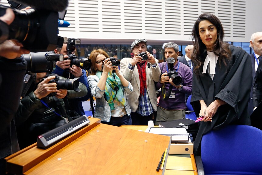 A woman in barrister's robes takes her place at a desk while photographers take her picture