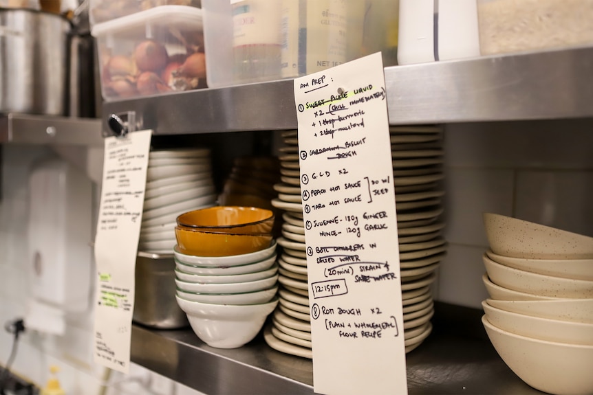 Two handwritten lists affixed to a commercial kitchen shelf with bowls on shelf