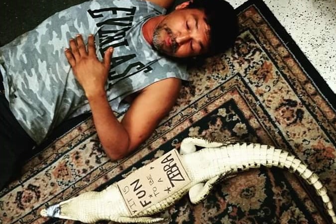 An Indigenous man lays next to a small croc with a sign saying its fun to be a zebra on its stomach