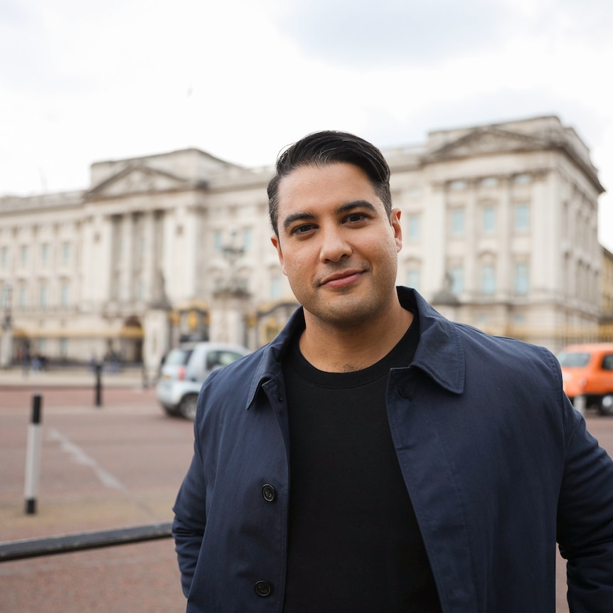 Marc smiles and looks to the camera as he stands in front of Buckingham Palace on a grey day, while wearing a navy jacket.