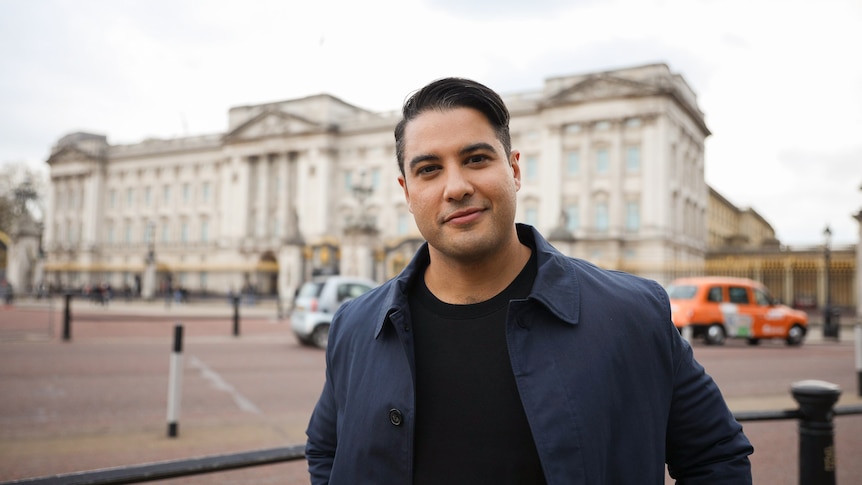 Marc smiles and looks to the camera as he stands in front of Buckingham Palace on a grey day, while wearing a navy jacket.