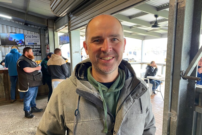 A man with a bald head, wearing a gray coat and a green jumper, smiles at the camera. 