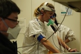 A woman on an exercise bike wearing breathing apparatus, with healthcare professionals on either side of her. 