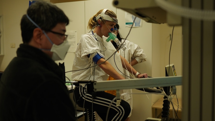 A woman on an exercise bike wearing breathing apparatus, with healthcare professionals on either side of her. 