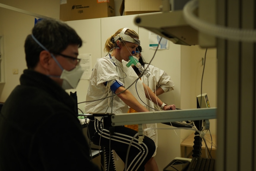 A woman on an exercise bike wearing breathing apparatus, with healthcare professionals on either side of her. 