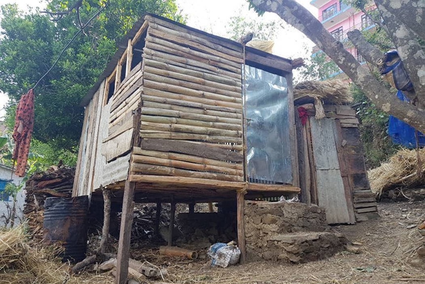 A rickety wooden hut sits outside a large multi story house.