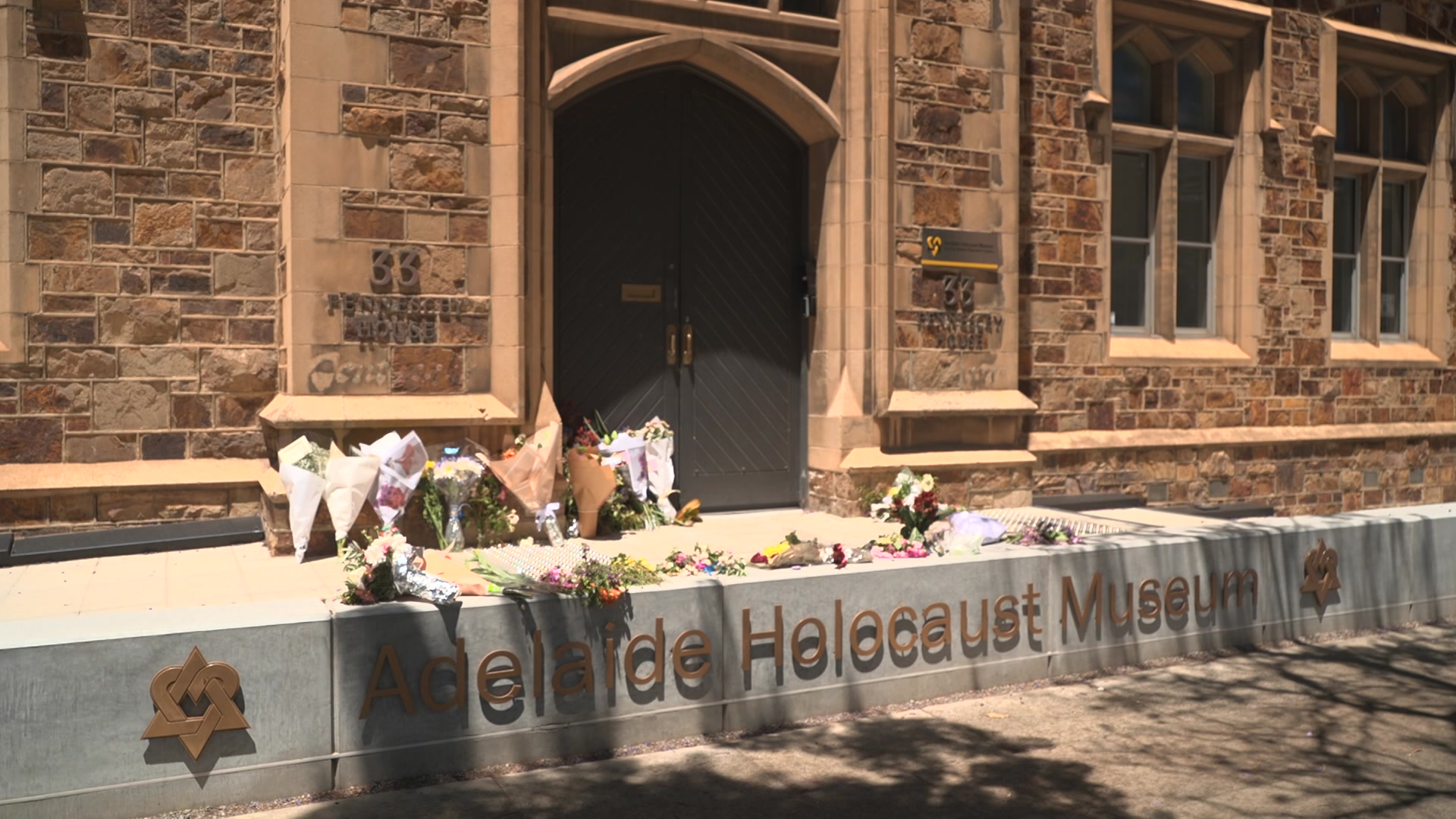 Ramos de flores frente a un antiguo edificio de ladrillo con grandes puertas. El letrero está en un escalón sobre el suelo.