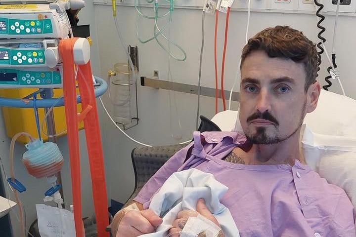 A man with brown hair, a mustache and arm tattoos lies in a hospital bed