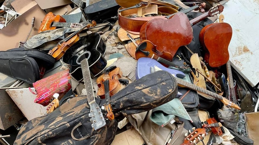 Musical instruments including guitars, violins, and a cello lie covered in mud in a rubbish heap.