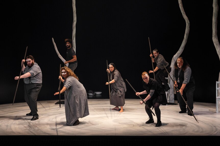 7 performers hold spears in a defensive stance in formation on stage wearing grey clothes and white face paint.