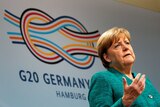 Angela Merkel stands in front of a G-20 banner and gestures with her hand.