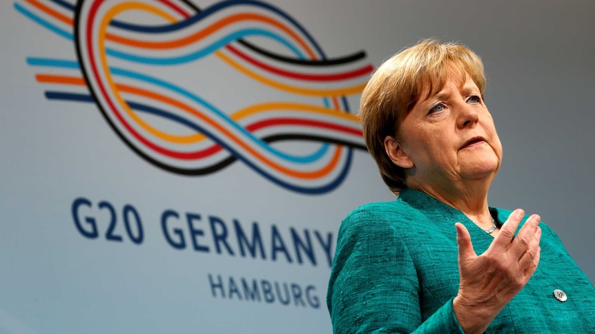 Angela Merkel stands in front of a G-20 banner and gestures with her hand.