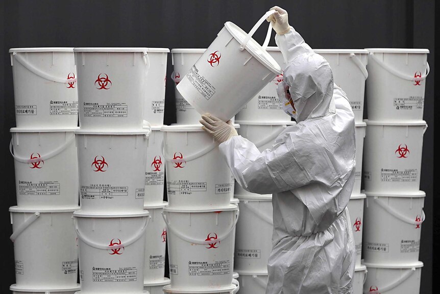 A worker in protective gear stacks a large wall of plastic buckets containing medical waste from coronavirus patients.