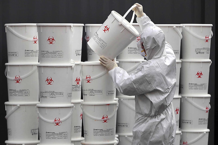 A worker in protective gear stacks a large wall of plastic buckets containing medical waste from coronavirus patients.