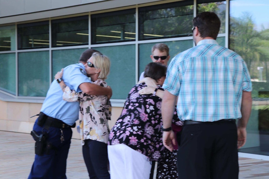 A group is standing around outside a building. A woman is hugging a police officer.