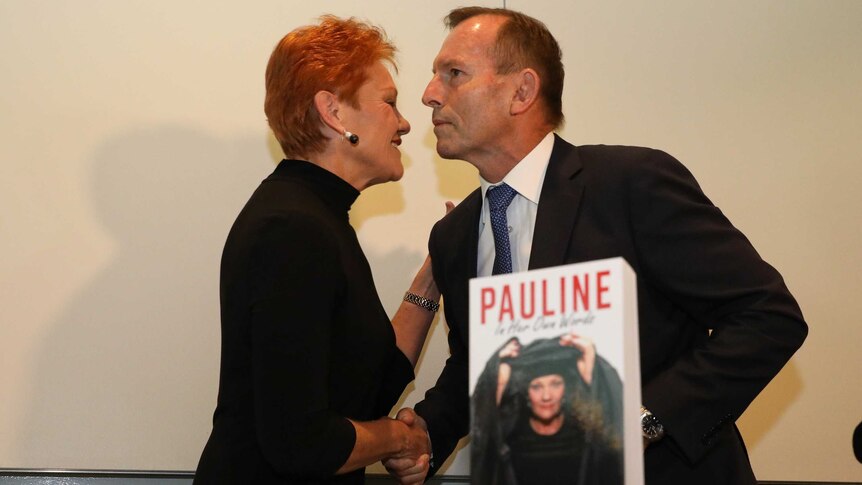 Tony Abbott and Pauline Hanson shake hands behind a copy of a the One Nation leader's book